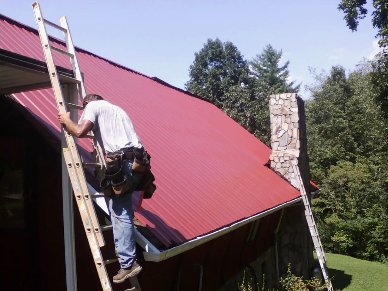 Roofer on a ladder beside a red metal roof and stone chimney — Soffit & Fascia Repair by Roofing Brothers in Hungerford